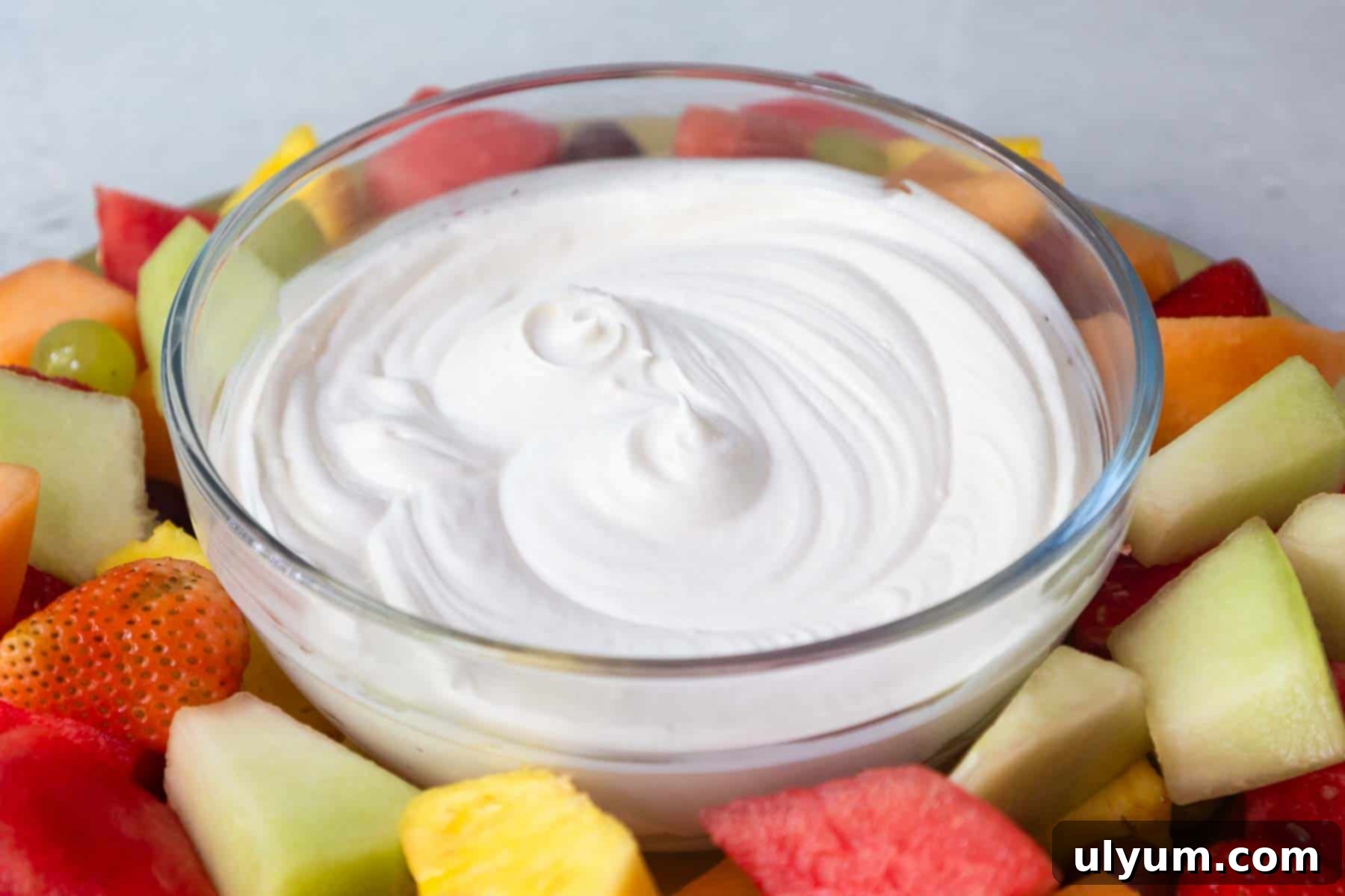 A glass bowl of smooth, swirled white fruit dip in the center of a vibrant assortment of cut fruit pieces arranged on a platter.