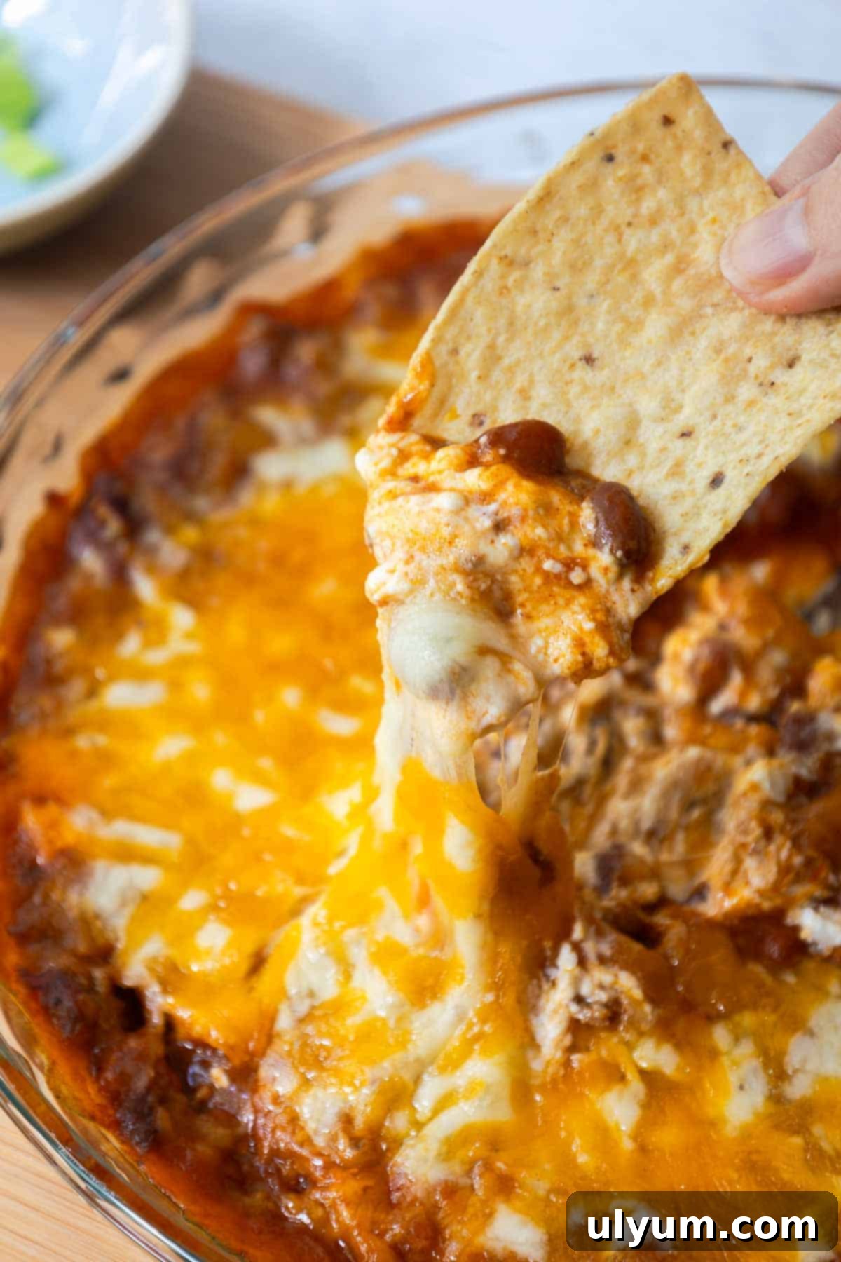 Tortilla chip being dipped into a hot, cheesy chili dip, with gooey layers of beans, cheese, and creamy filling stretching upward.