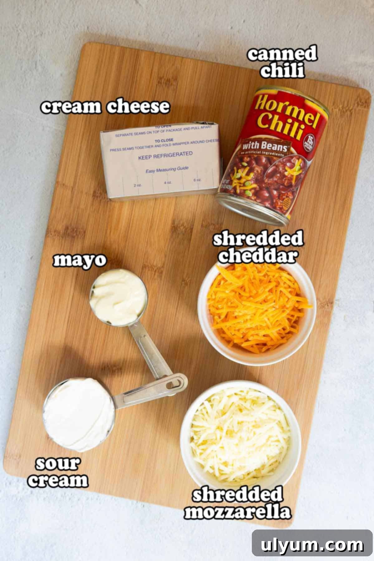 Overhead view of labeled chili cheese dip ingredients on a wooden cutting board, including canned chili, cream cheese, mayo, sour cream, shredded cheddar, and shredded mozzarella.