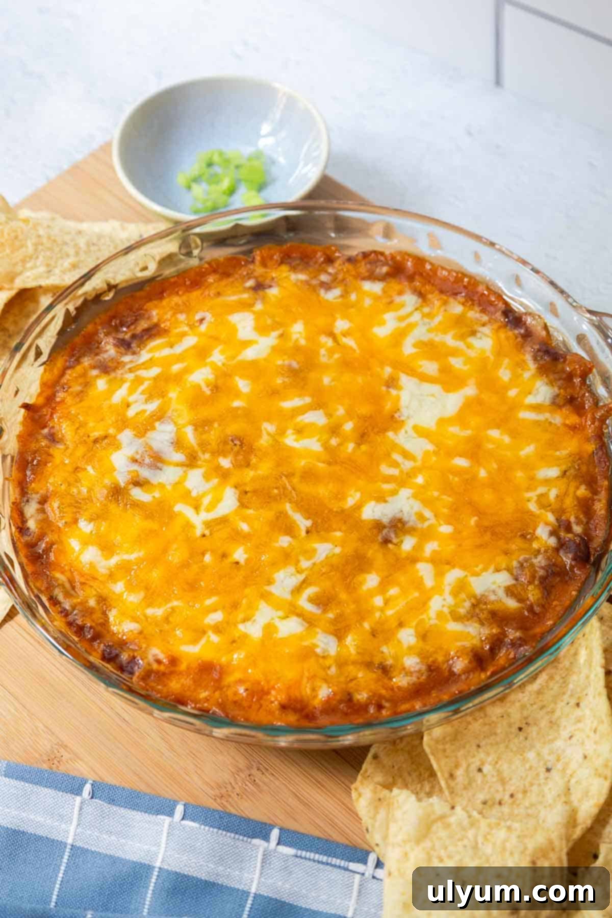 A glass pie dish filled with hot chili cheese dip, topped with melted cheddar and mozzarella, surrounded by tortilla chips and a small bowl of green onions.