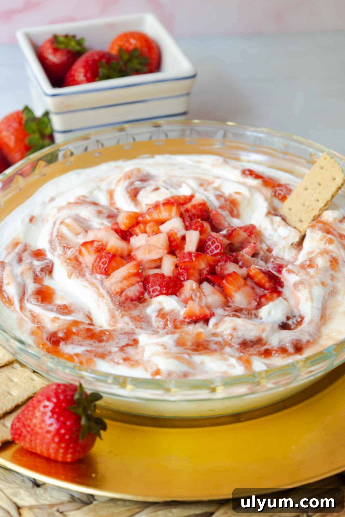 Glass pie plate filled with swirled strawberry and cream cheese dip, topped with fresh diced strawberries and a graham cracker for dipping.