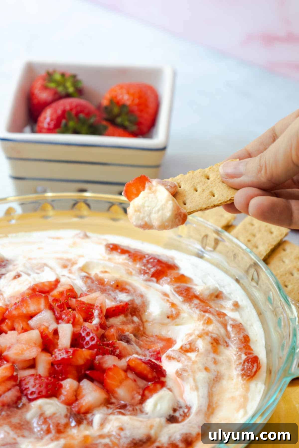 Close-up of a hand dipping a graham cracker into a creamy strawberry dessert, topped with fresh strawberry chunks.