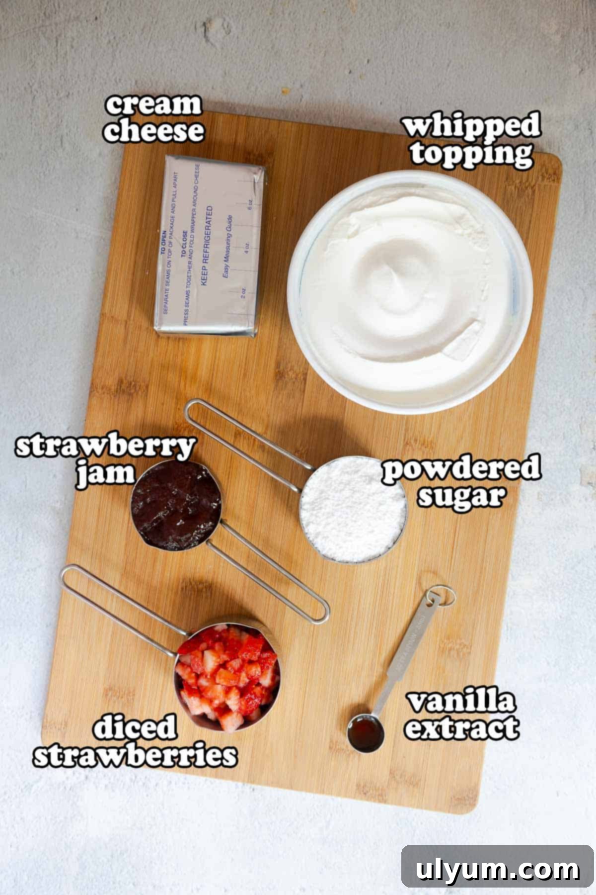 Ingredients for strawberry cheesecake dip on a wooden board, labeled as cream cheese, whipped topping, powdered sugar, strawberry jam, diced strawberries, and vanilla extract.