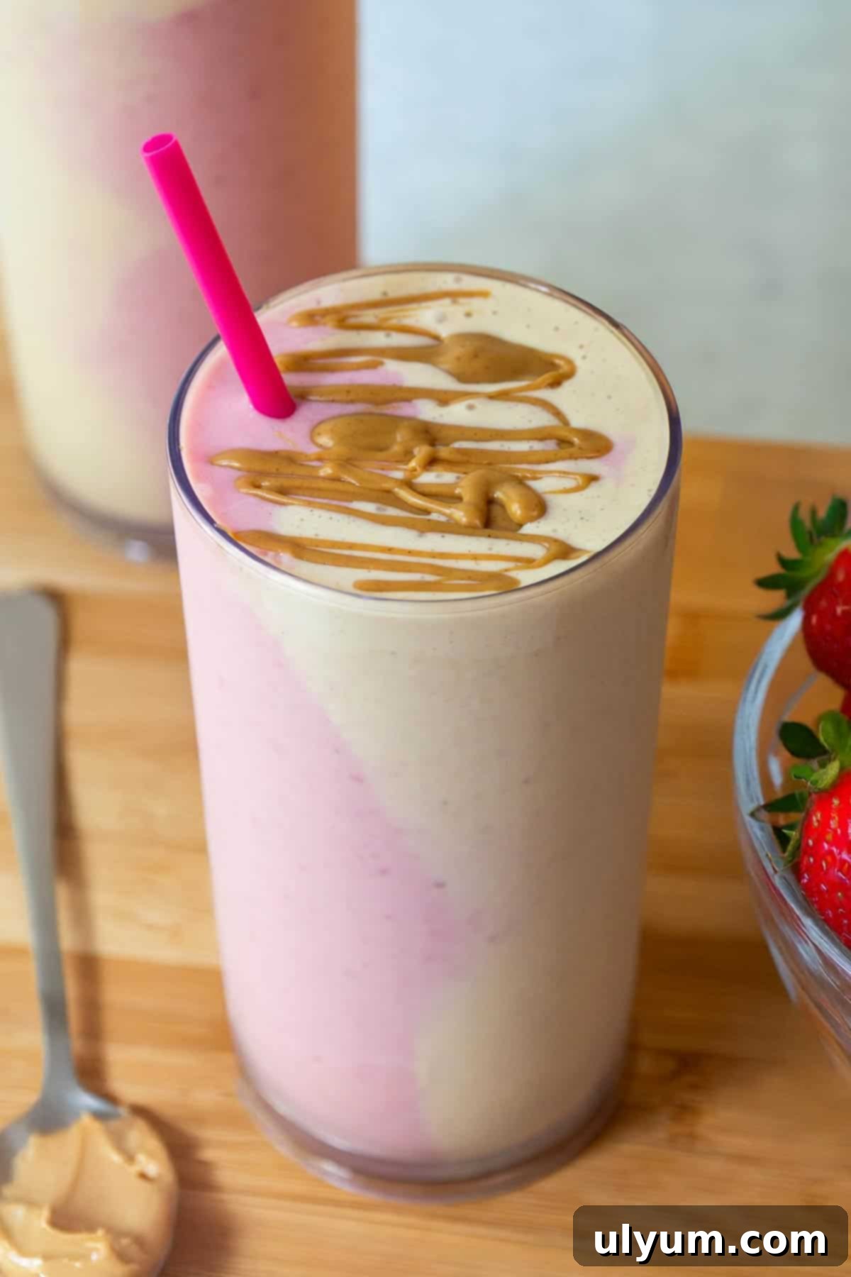 A tall glass of peanut butter and jelly smoothie with pink and beige side-by-side in the glass, topped with a drizzle of peanut butter.