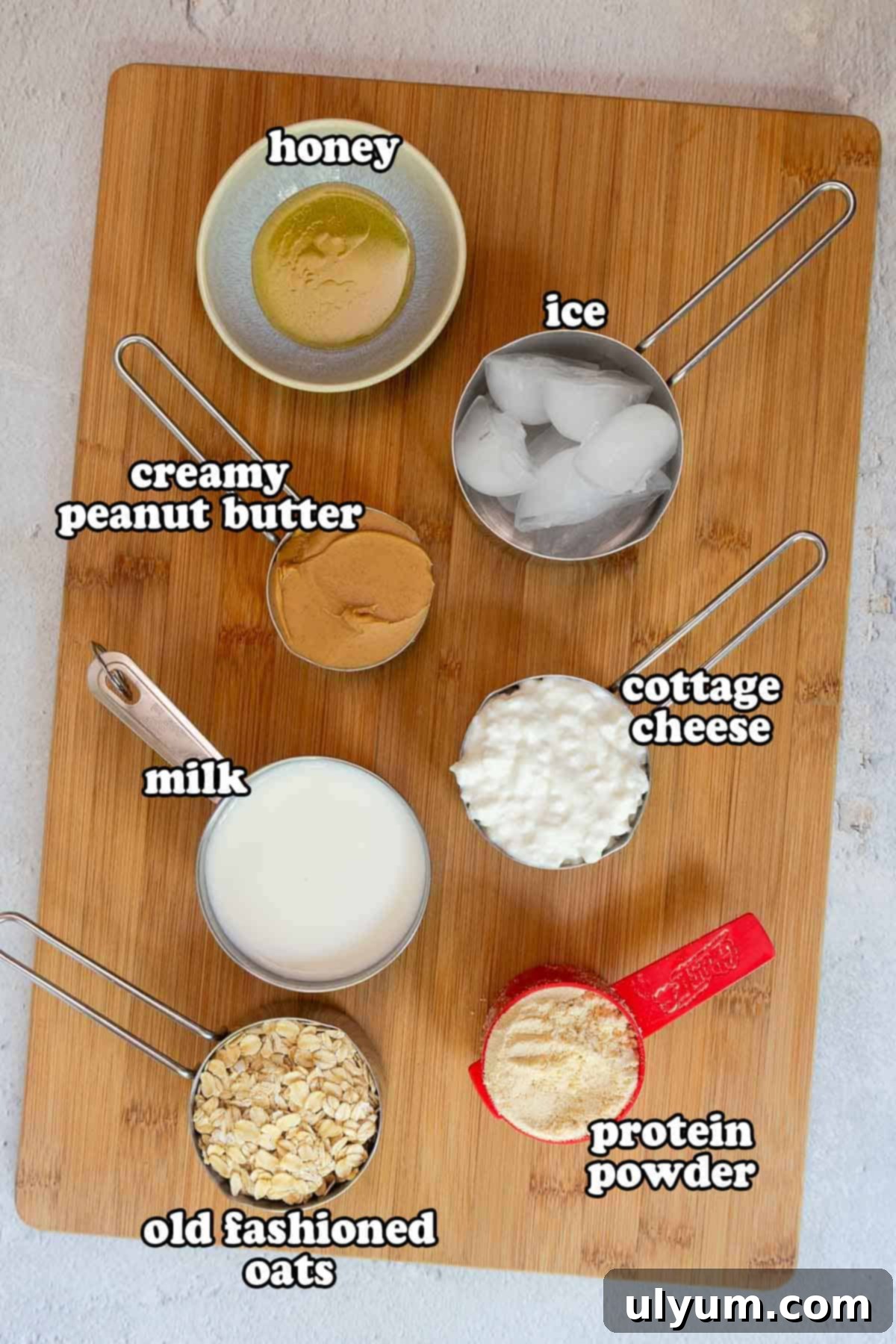 A wooden cutting board topped with smoothie ingredients labeled: honey, ice, creamy peanut butter, milk, old fashioned oats, cottage cheese, and protein powder.
