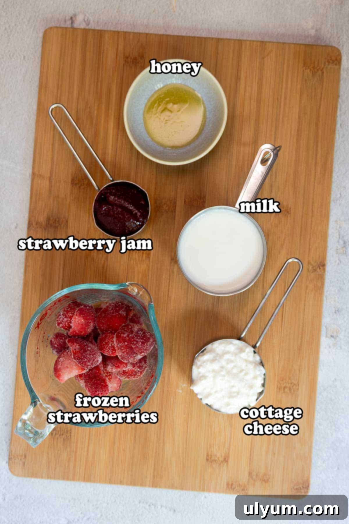 Ingredients for the jelly smoothie on a wooden cutting board, labeled: honey, milk, strawberry jam, frozen strawberries, and cottage cheese.