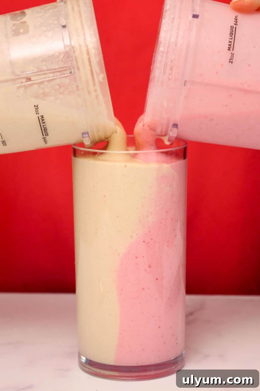 Two blender cups simultaneously pouring the peanut butter and jelly smoothies side-by-side into a single glass, creating a swirled effect.