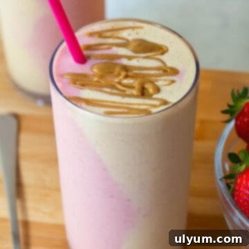 A tall glass of peanut butter and jelly smoothie with pink and beige side-by-side in the glass, topped with peanut butter drizzle.