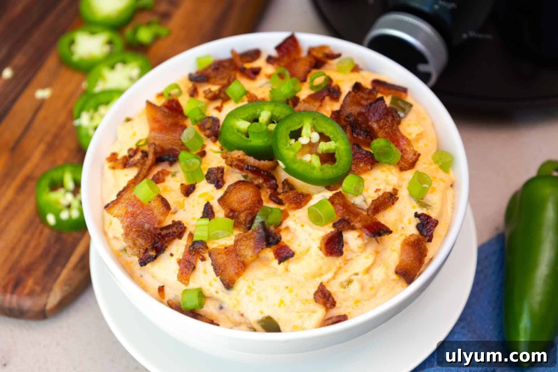 Close-up of jalapeño popper dip in a white bowl, garnished with chopped bacon, green onions, and fresh jalapeño slices, with a Crockpot in the background.