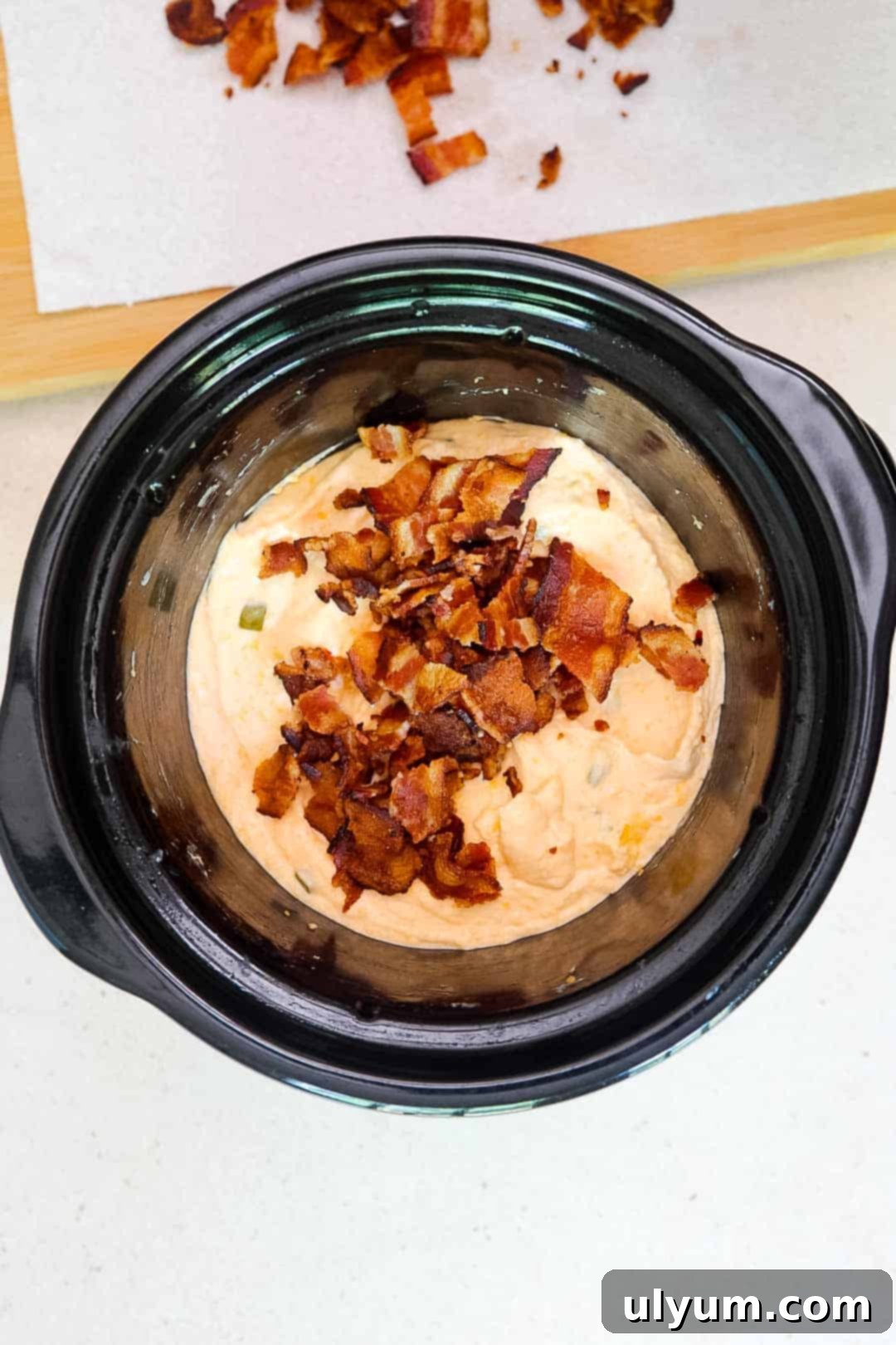 Slow cooker with cooked jalapeño popper dip topped with a generous amount of chopped bacon, before final mixing.