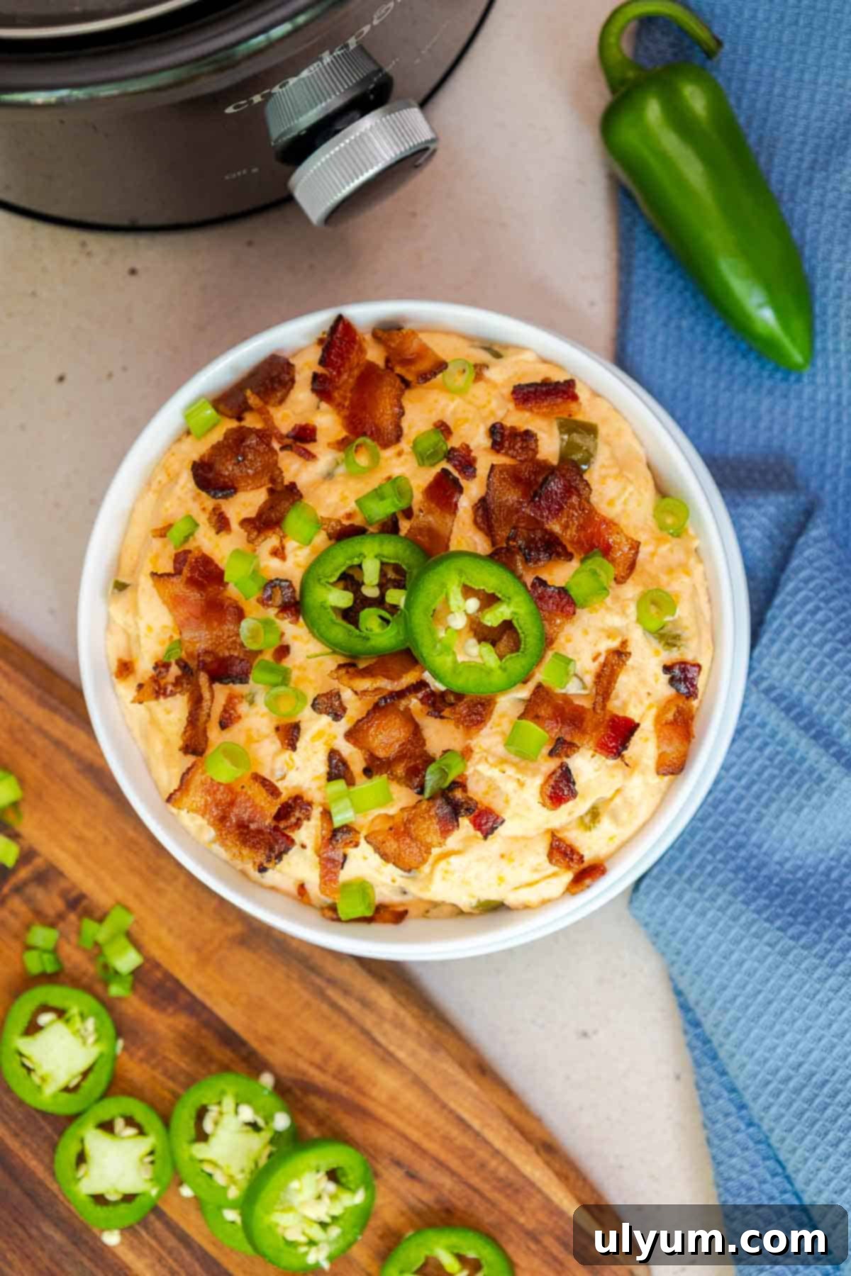 Overhead shot of finished crockpot jalapeño popper dip in a white bowl, garnished with sliced jalapeños, crispy bacon, and green onions, next to a green jalapeño and a Crockpot.