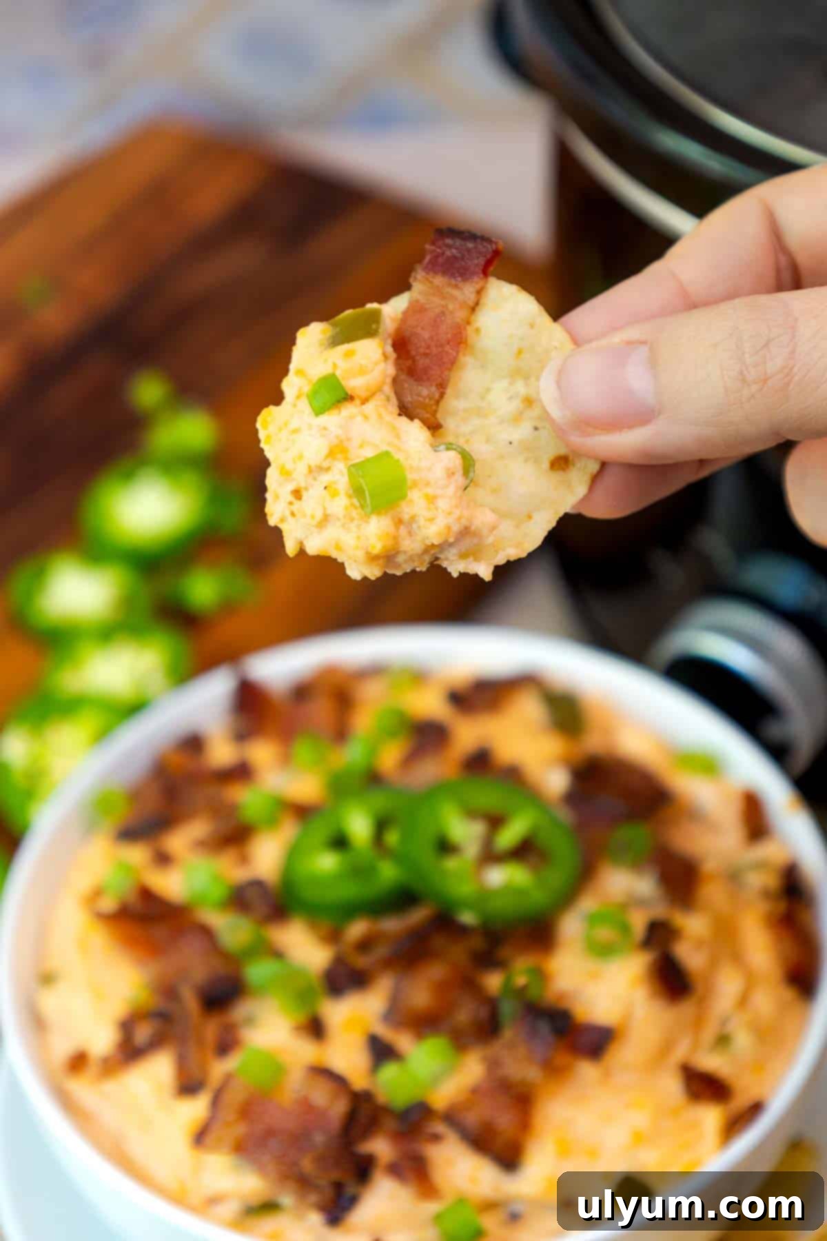 A hand holding a tortilla chip scooped with creamy jalapeño popper dip, showing melted cheese, jalapeños, bacon, and green onion bits.