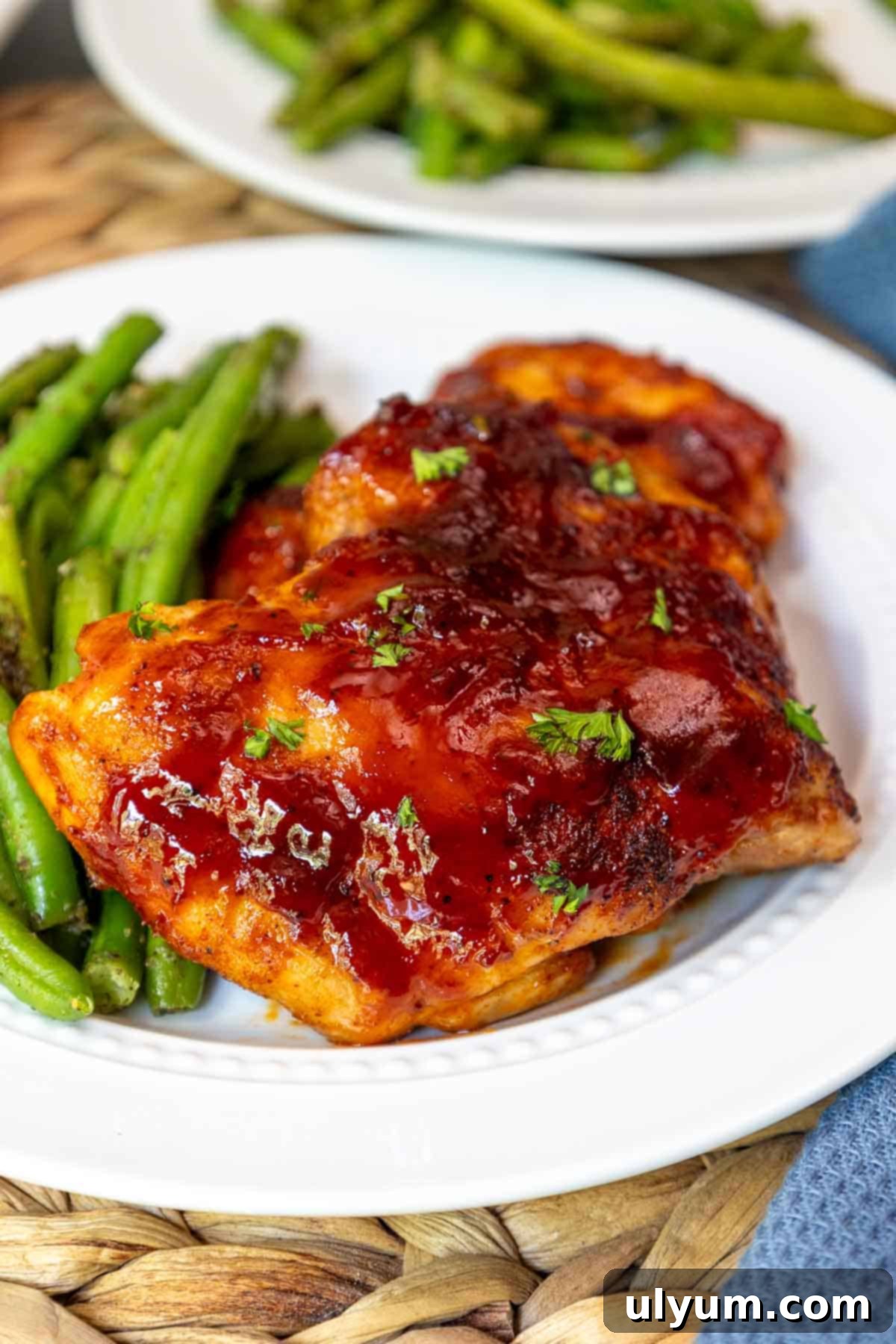 Mouthwatering Oven Baked BBQ Boneless Chicken Thighs 2 Close-up of oven baked BBQ boneless chicken thighs glazed with sauce and sprinkled with parsley, served with green beans on a white plate.