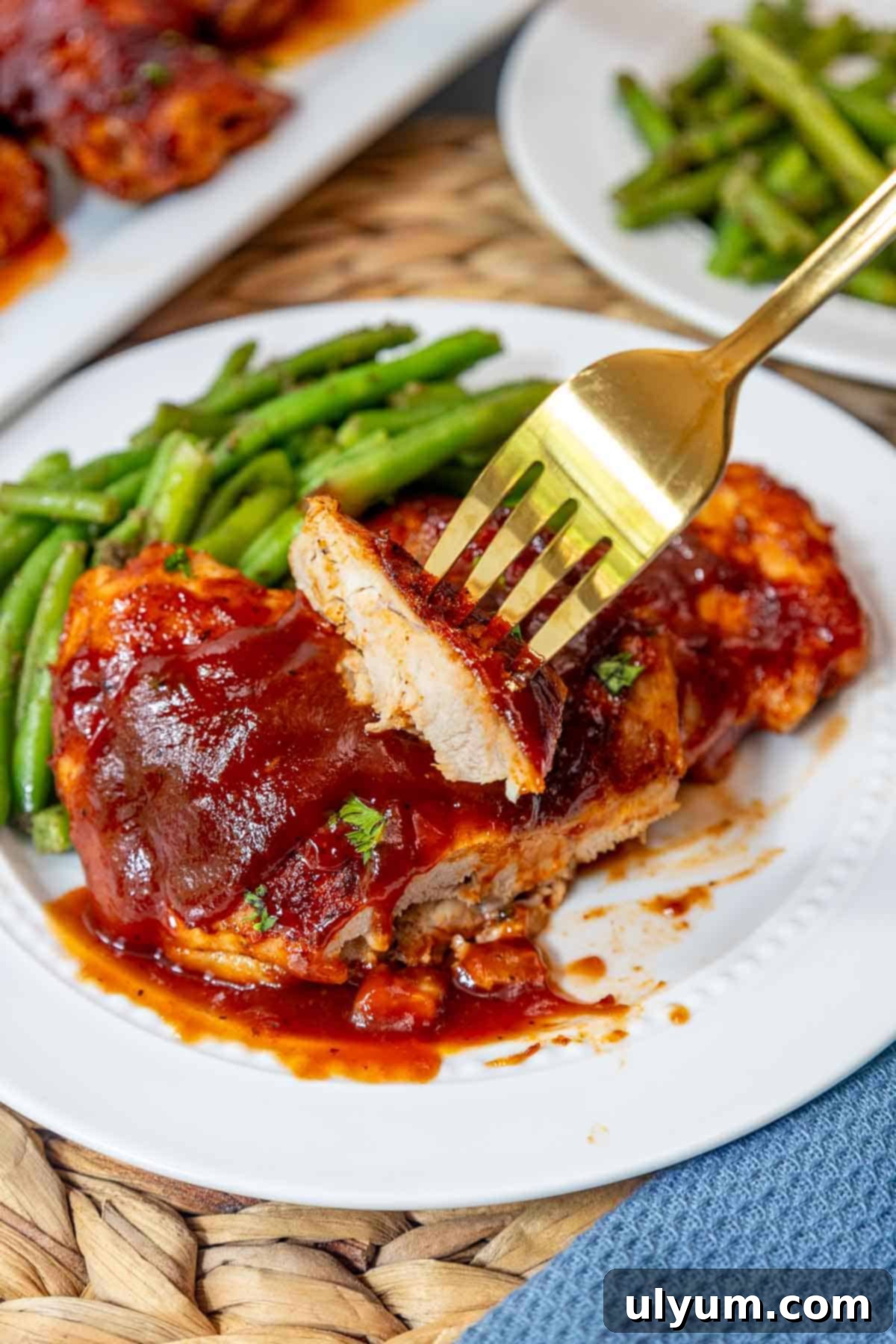 Mouthwatering Oven Baked BBQ Boneless Chicken Thighs 11 A bite of tender BBQ chicken thigh being lifted by a fork, revealing the juicy inside of the oven baked boneless meat next to green beans.
