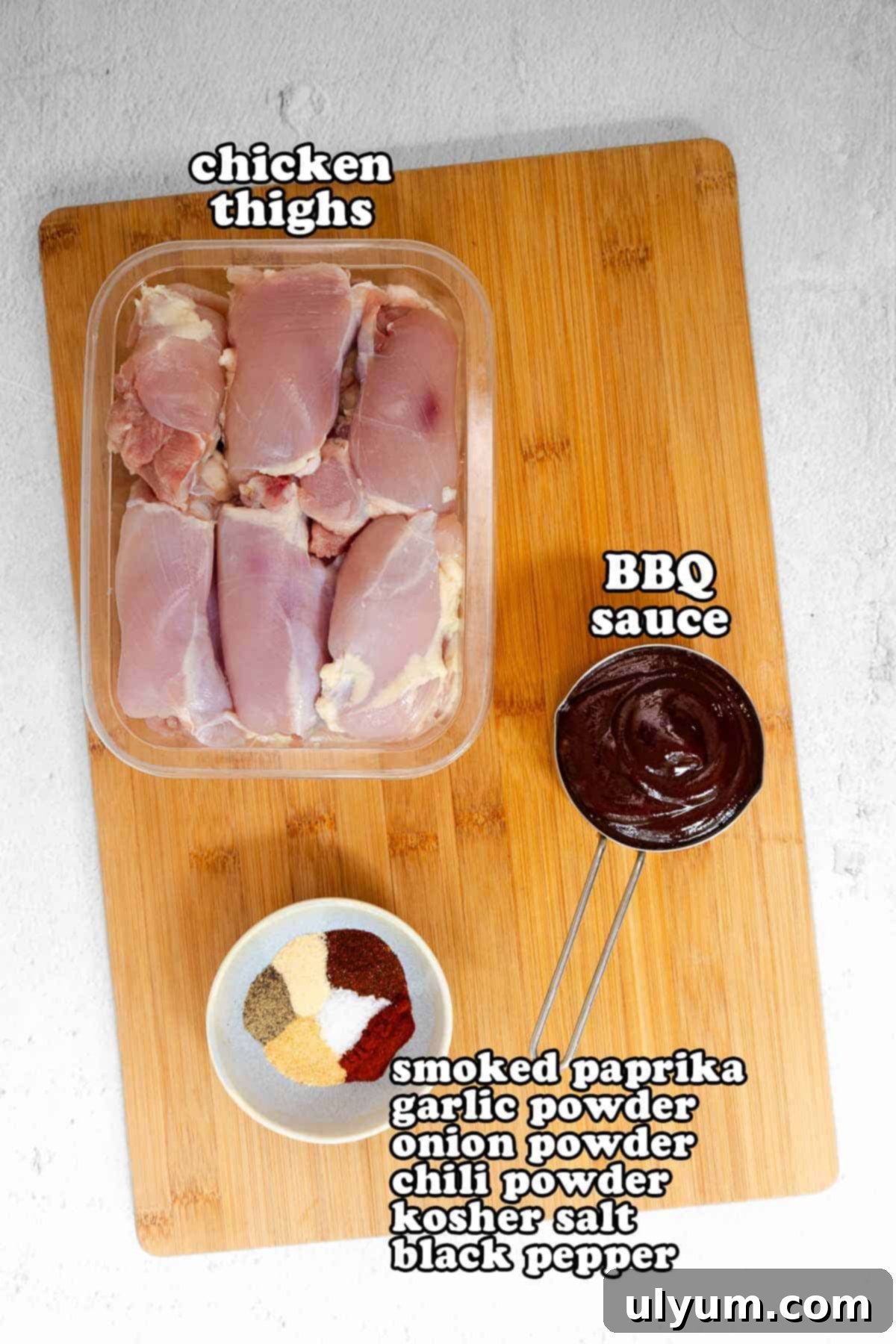 Mouthwatering Oven Baked BBQ Boneless Chicken Thighs 3 Flat lay of raw ingredients on a wooden cutting board including boneless chicken thighs, a bowl of BBQ sauce, and a spice blend labeled with smoked paprika, garlic powder, onion powder, chili powder, kosher salt, and black pepper.