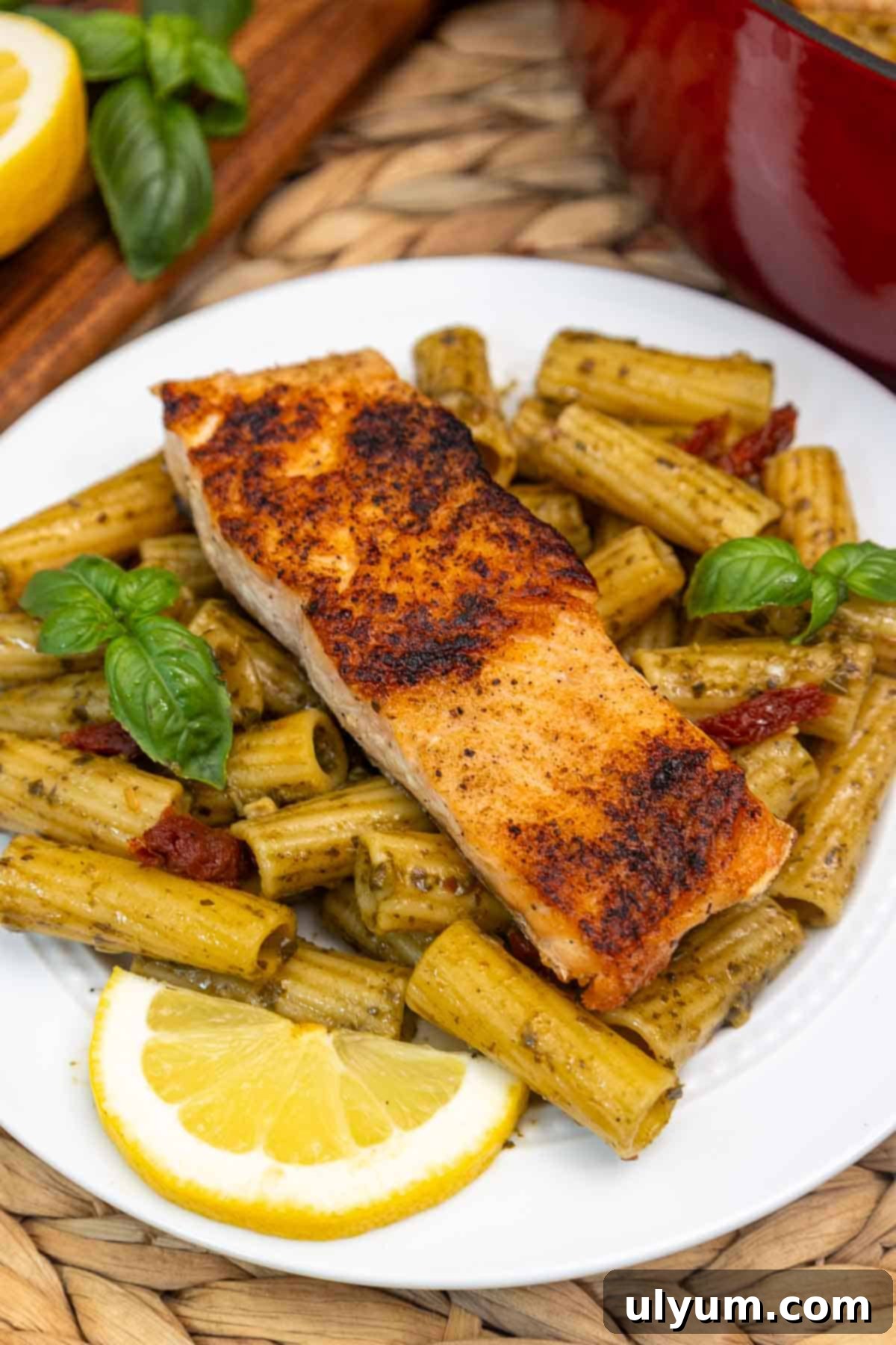 Plate of salmon pesto pasta with a seared salmon fillet on top of rigatoni tossed in pesto sauce, sun-dried tomatoes, and garnished with fresh basil and a lemon wedge.