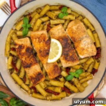Pan of salmon pesto pasta with four seared salmon fillets on top of rigatoni tossed in pesto sauce, sun-dried tomatoes, and garnished with fresh basil and a lemon wedge.
