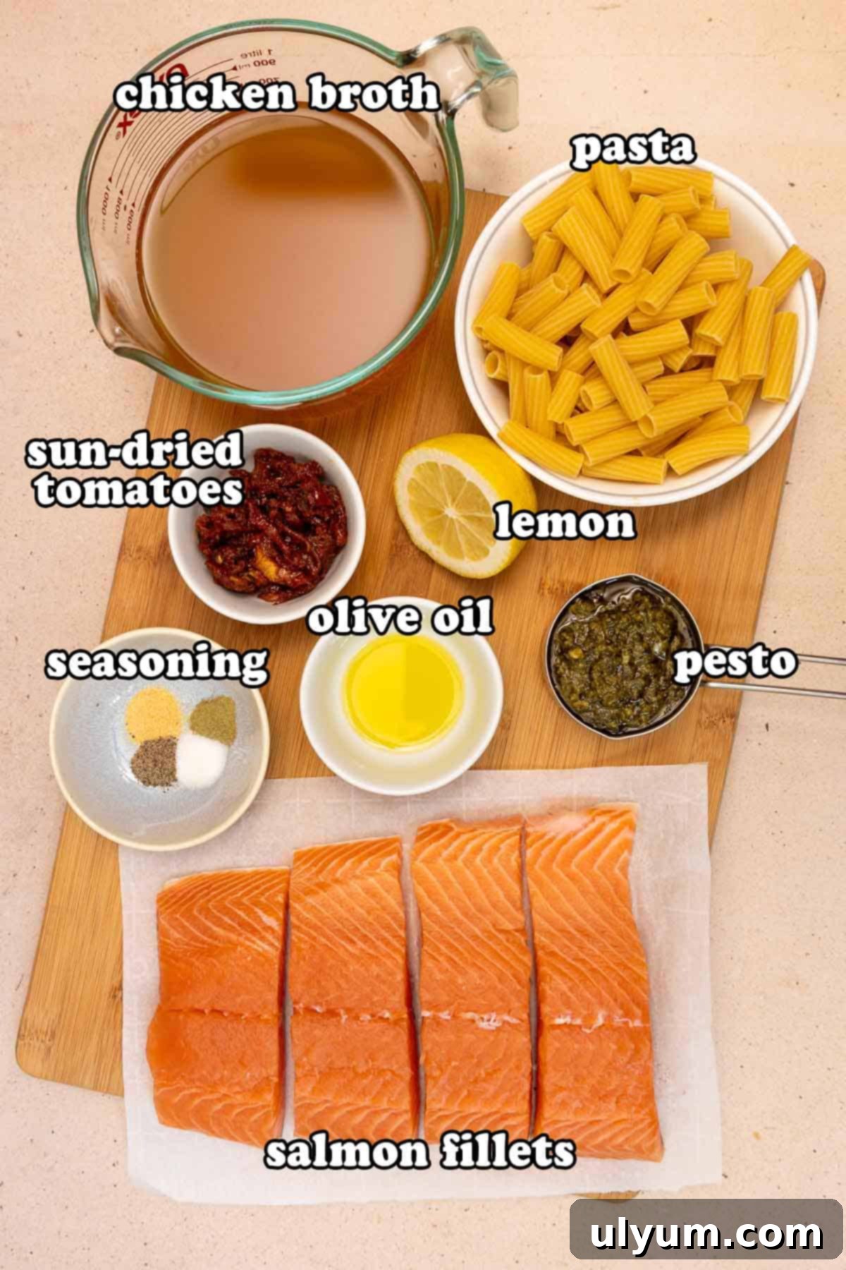 Salmon pesto pasta ingredients on a cutting board, including raw salmon fillets, dry rigatoni pasta, chicken broth, sun-dried tomatoes, lemon, olive oil, pesto, and small bowls of seasonings.