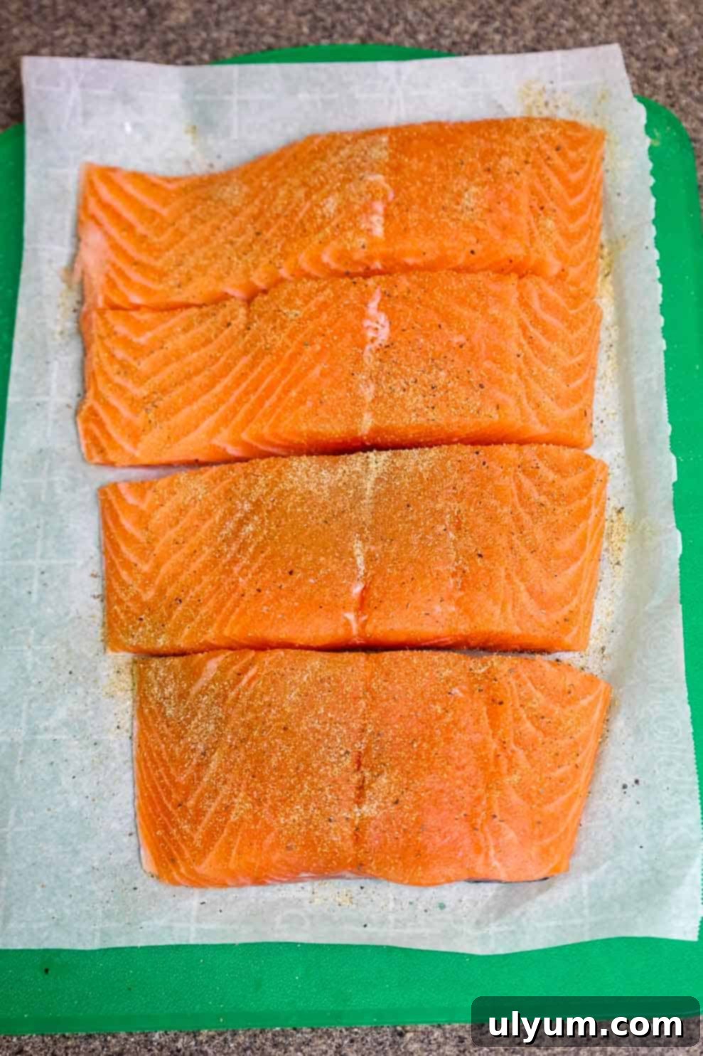 Four raw salmon fillets evenly seasoned and arranged on parchment paper.