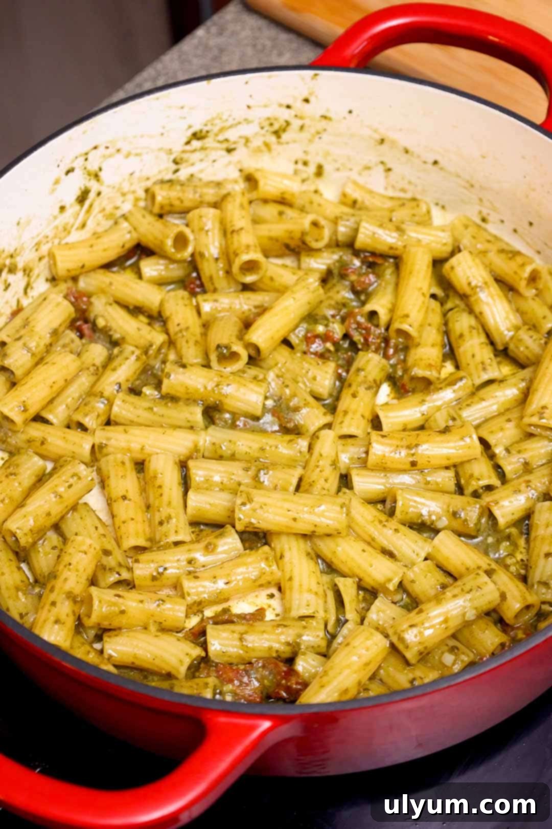 Cooked rigatoni pasta in a Dutch oven, coated in pesto sauce and mixed with bits of sun-dried tomatoes.