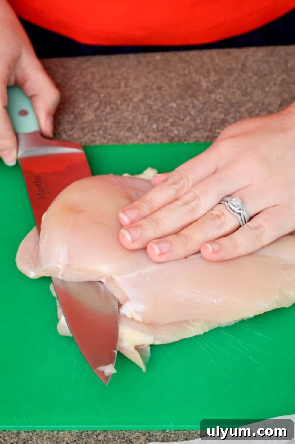 A person slicing a raw chicken breast in half horizontally on a cutting board with a large chef’s knife.