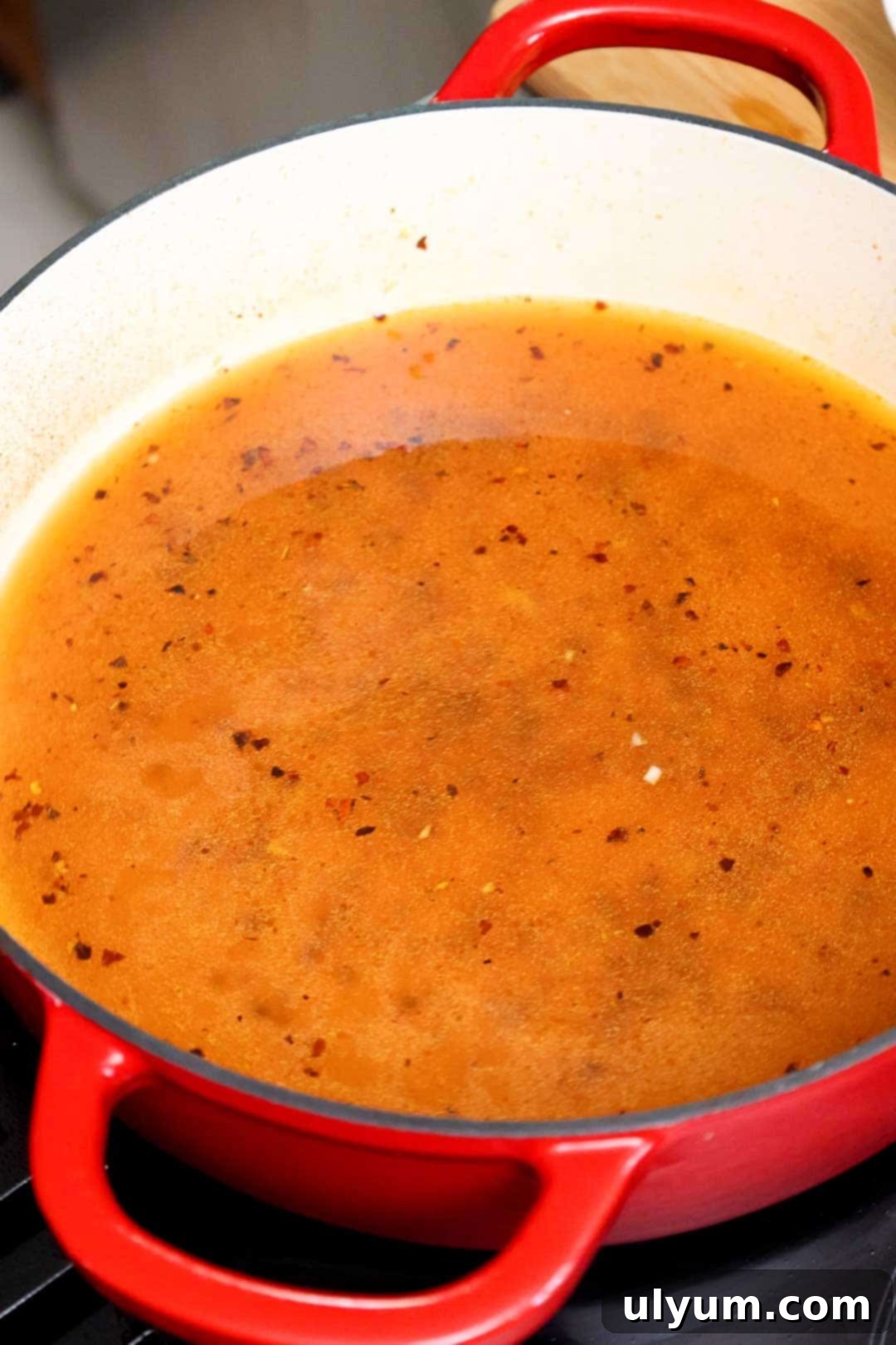 Chicken broth simmering in a red Dutch oven, speckled with red pepper flakes and cooking residue.