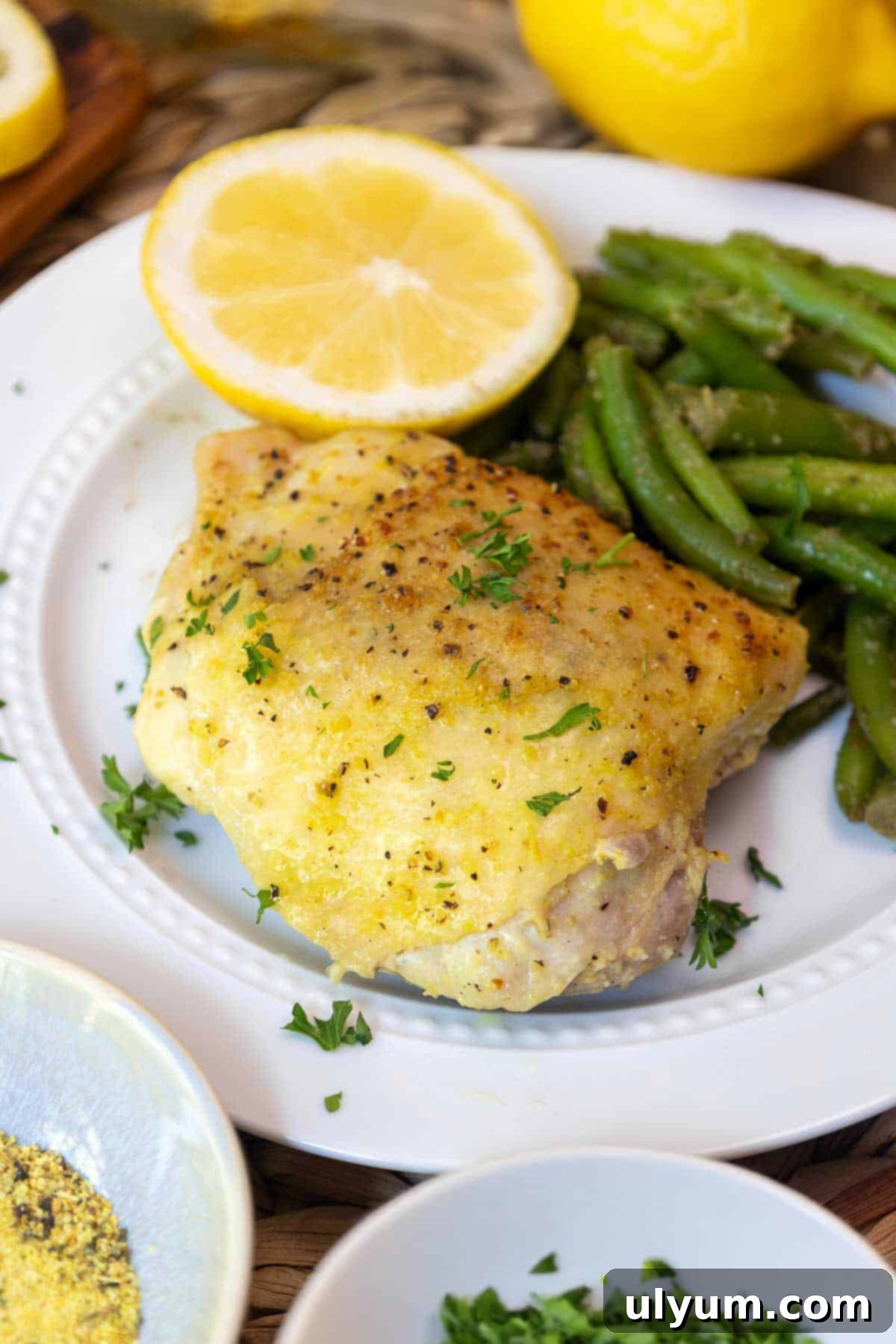 Golden Lemon Pepper Chicken Thighs 2 A plated serving of baked lemon pepper chicken thigh garnished with parsley, served with sautéed green beans and a halved lemon on a white plate.