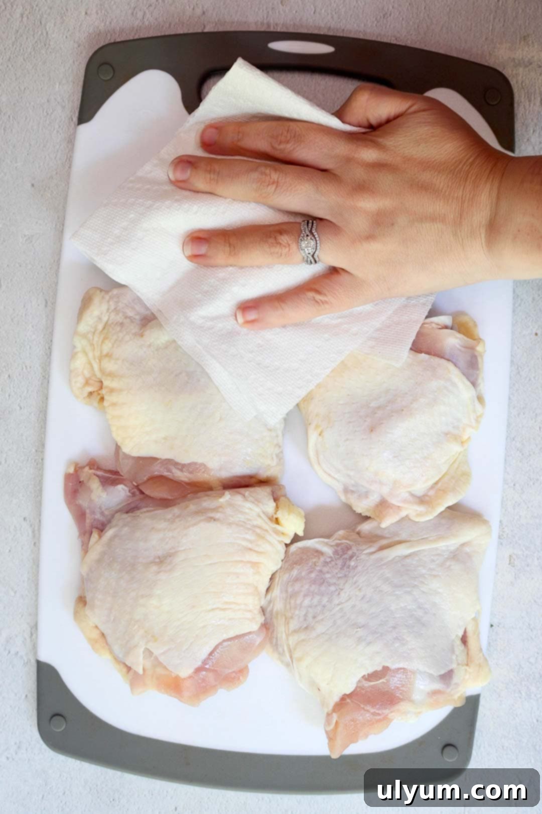 Golden Lemon Pepper Chicken Thighs 4 A hand using a paper towel to pat dry raw chicken thighs on a white cutting board.