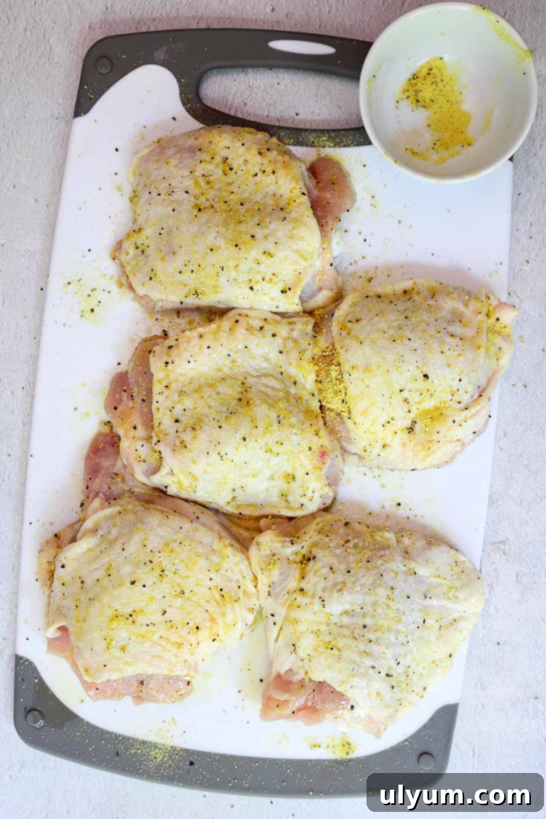 Golden Lemon Pepper Chicken Thighs 6 Raw chicken thighs thoroughly seasoned with lemon pepper blend on a cutting board, with visible specks of black pepper and yellow seasoning.