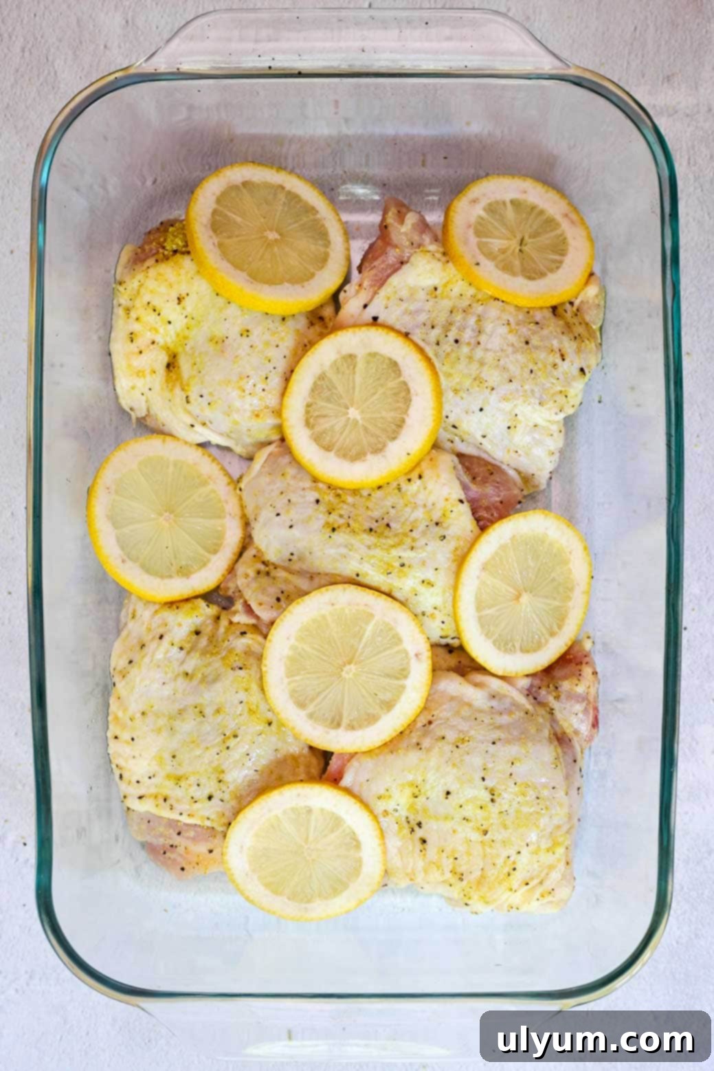 Golden Lemon Pepper Chicken Thighs 7 Seasoned raw chicken thighs arranged in a glass baking dish, topped with lemon slices ready for baking.