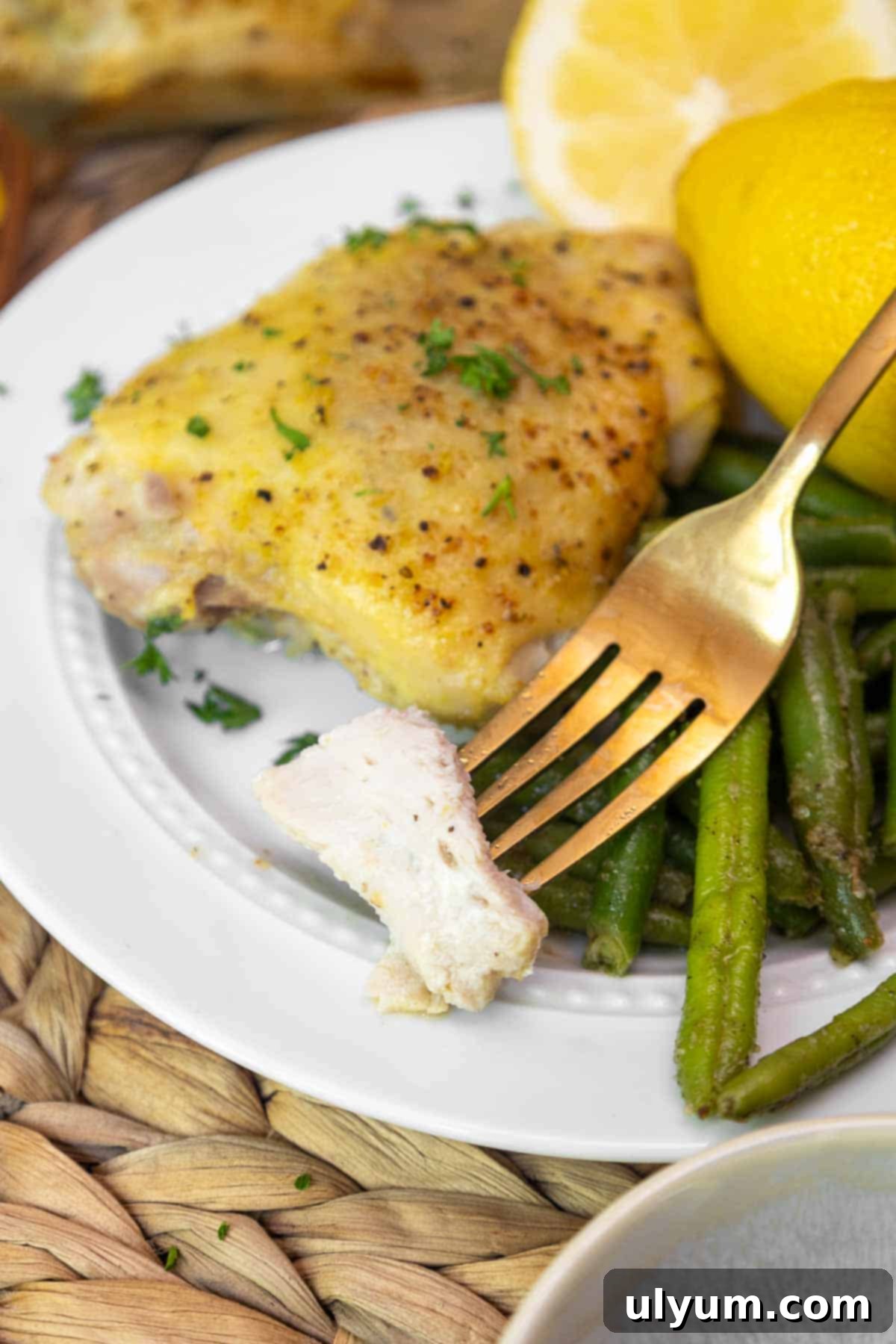 Golden Lemon Pepper Chicken Thighs 9 A fork holding a bite-sized piece of juicy baked chicken thigh above a plate of lemon pepper chicken, green beans, and fresh lemon.