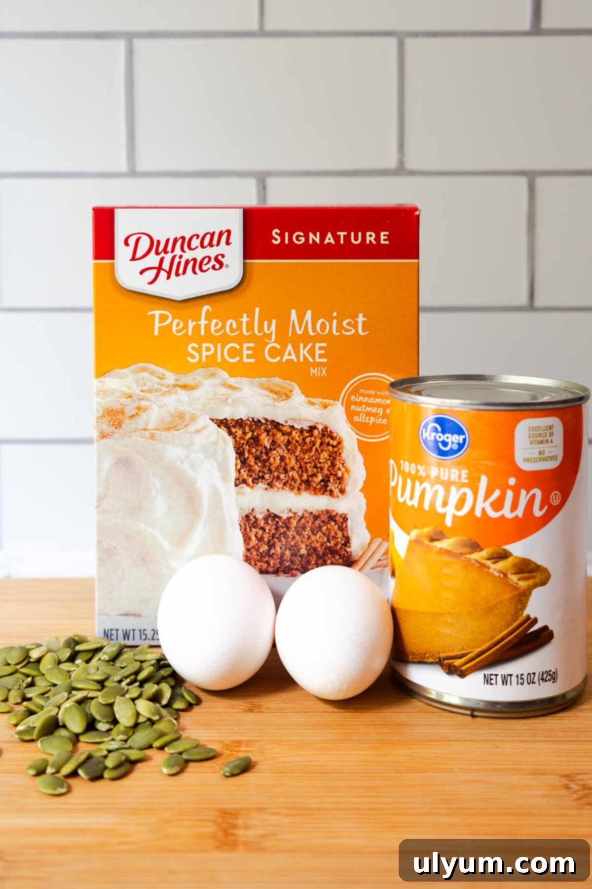 Pumpkin Spice Cake Loaf 3 Ingredients for pumpkin bread with spice cake mix arranged on a wooden cutting board, including Duncan Hines spice cake mix, a can of Kroger pure pumpkin, two eggs, and a handful of raw pepitas.