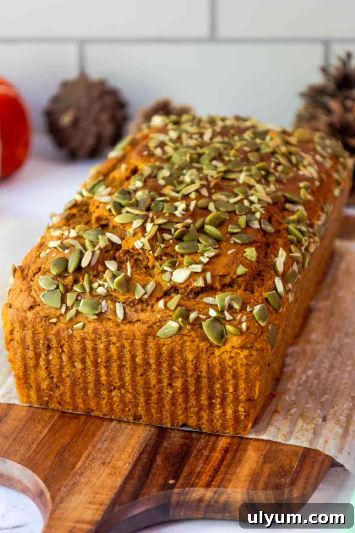 Pumpkin Spice Cake Loaf 8 A whole loaf of golden pumpkin bread with a domed top, generously sprinkled with pumpkin seeds, resting on a wooden cutting board.