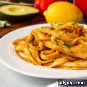 A tempting image of creamy avocado bell pepper pasta served on a plate, garnished with fresh basil. Fresh avocado, red bell pepper, and lemon are artfully arranged in the background.