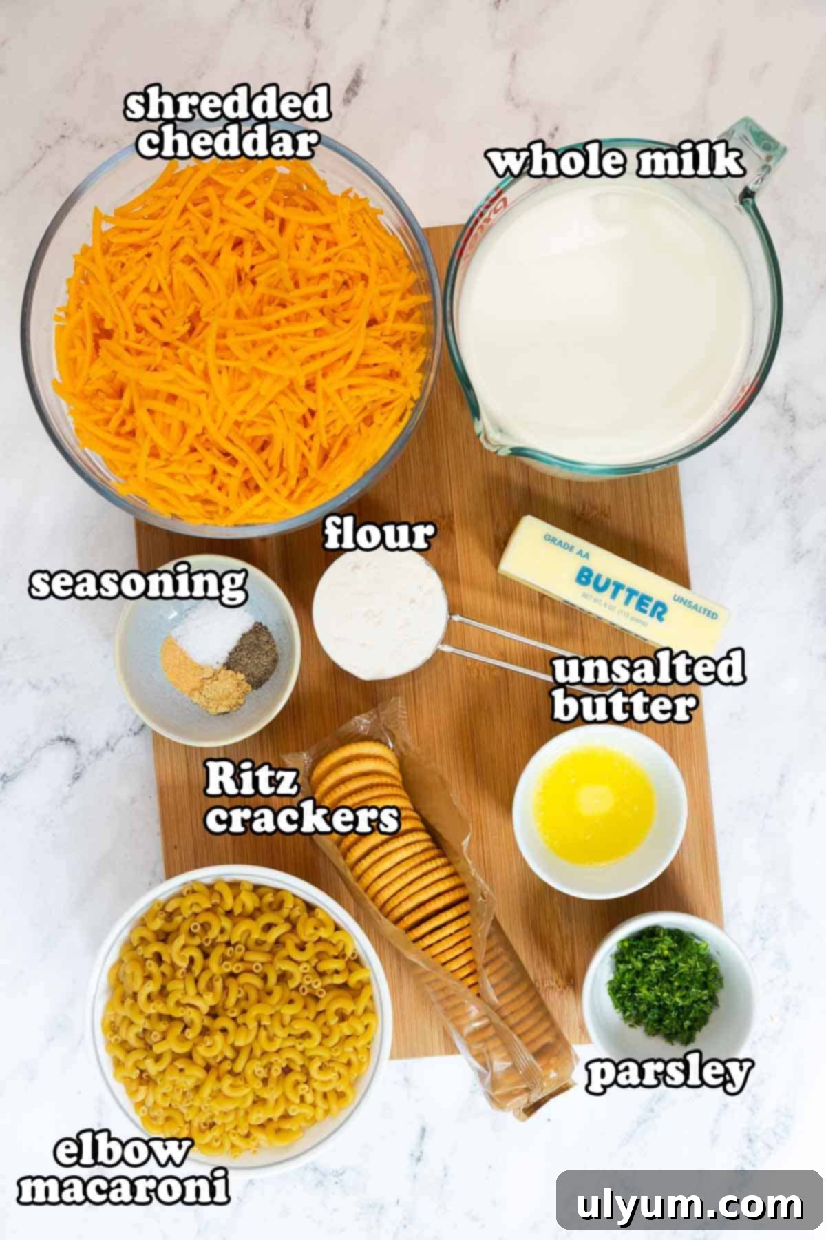 Ingredients for old fashioned baked mac and cheese with Ritz crackers displayed on a wooden board, including shredded cheddar, whole milk, unsalted butter, flour, elbow macaroni, Ritz crackers, seasoning, and parsley.