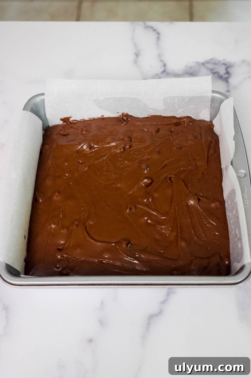 Brownie batter spread evenly in a parchment-lined square baking pan, ready to be baked.