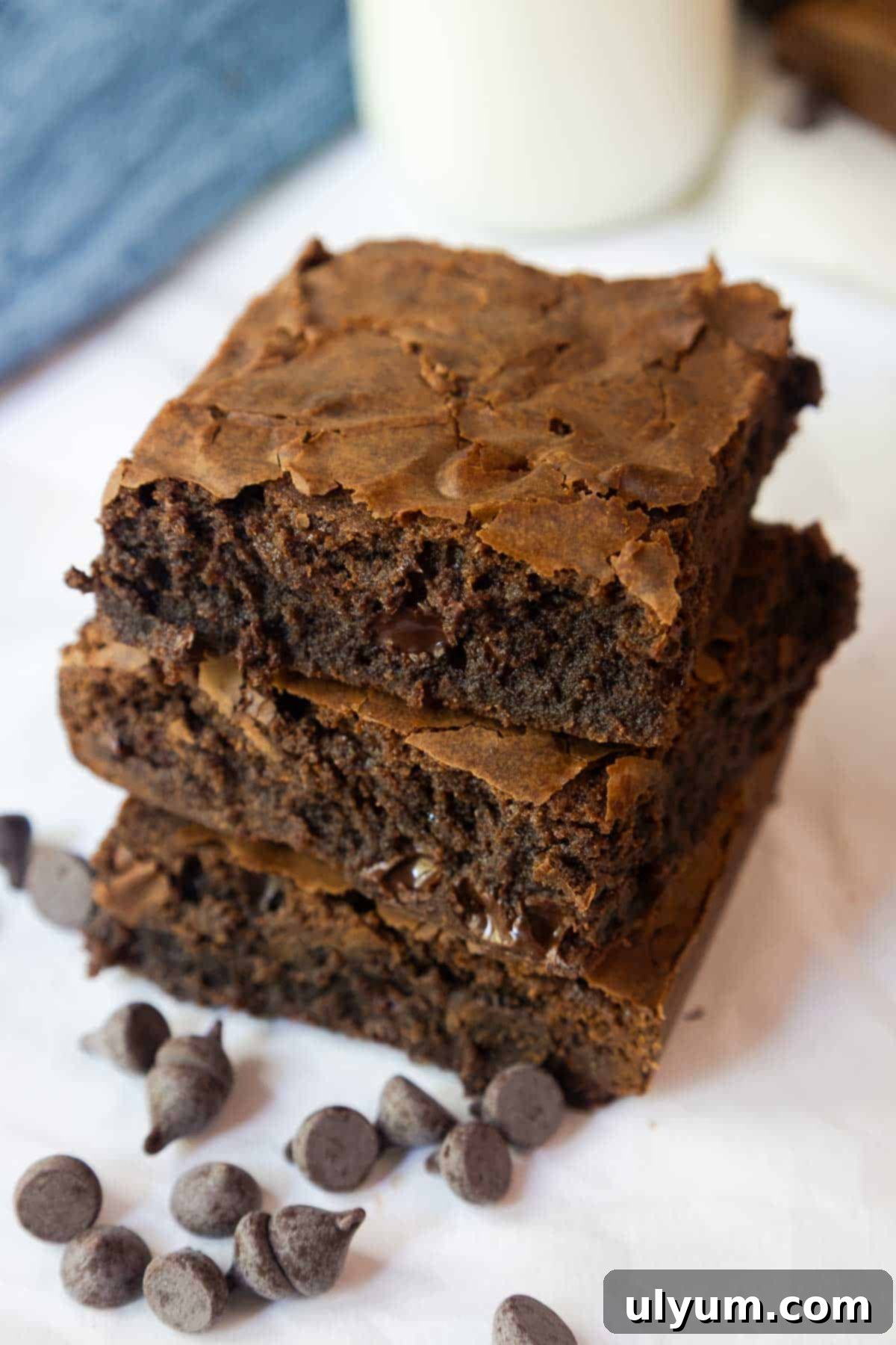 Stack of brownies next to chocolate chips and a glass of milk, highlighting the moist interior and crisp top layer.