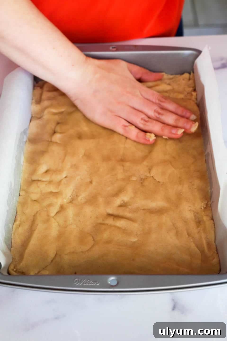 Dough being pressed by hand evenly into a lined baking pan.