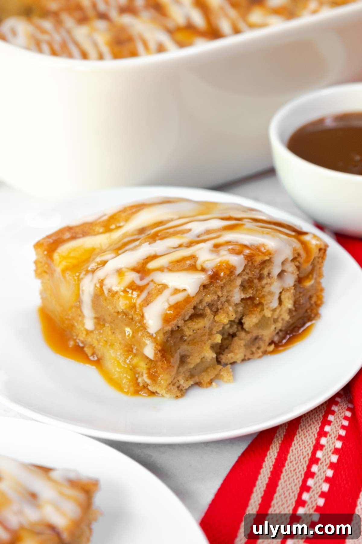 A moist slice of apple cake drizzled with vanilla glaze and caramel sauce, served on a small plate.