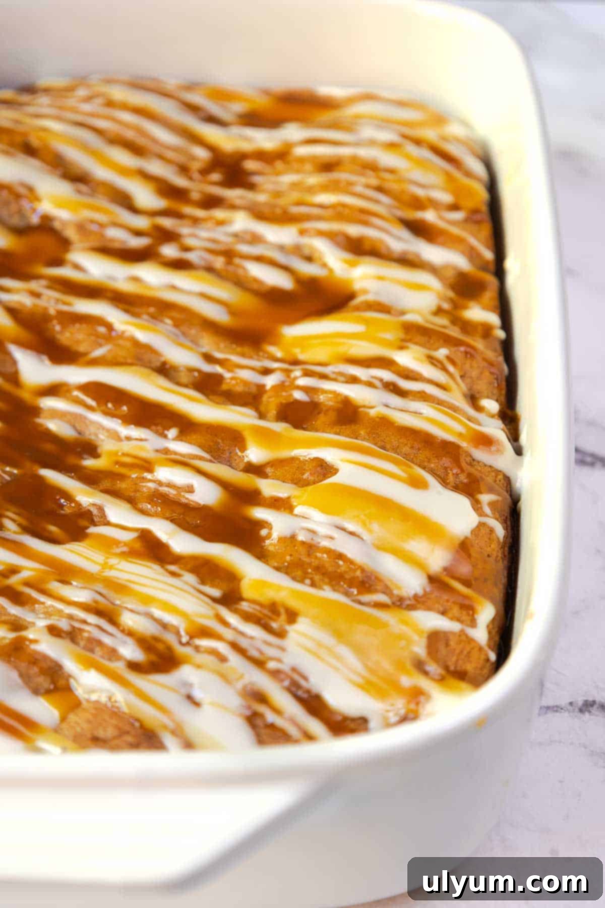 Overhead view of a golden-brown apple cake covered in zigzag drizzles of powdered sugar glaze and caramel sauce in a 9 by 13 inch baking dish.