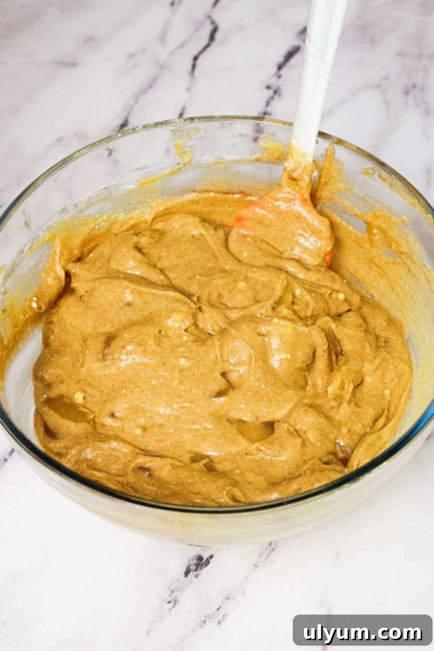 Thick, cinnamon-speckled apple cake batter in a glass mixing bowl with a spatula, ready to be poured into a baking dish.