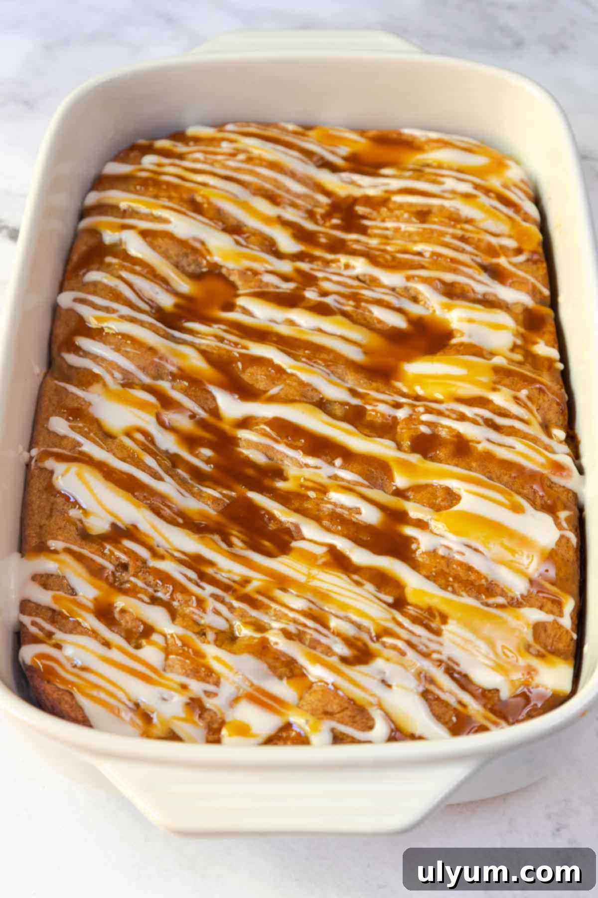 The finished apple cake in a 9 by 13 inch baking dish, topped with a generous drizzle of both white glaze and caramel sauce.