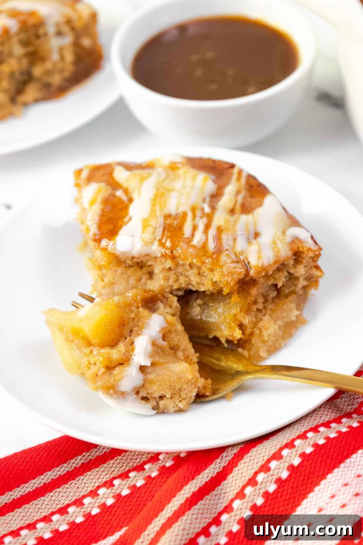 A bite of apple cake on a fork reveals tender apple pieces inside the spiced cake, with glaze and caramel drizzle on top.