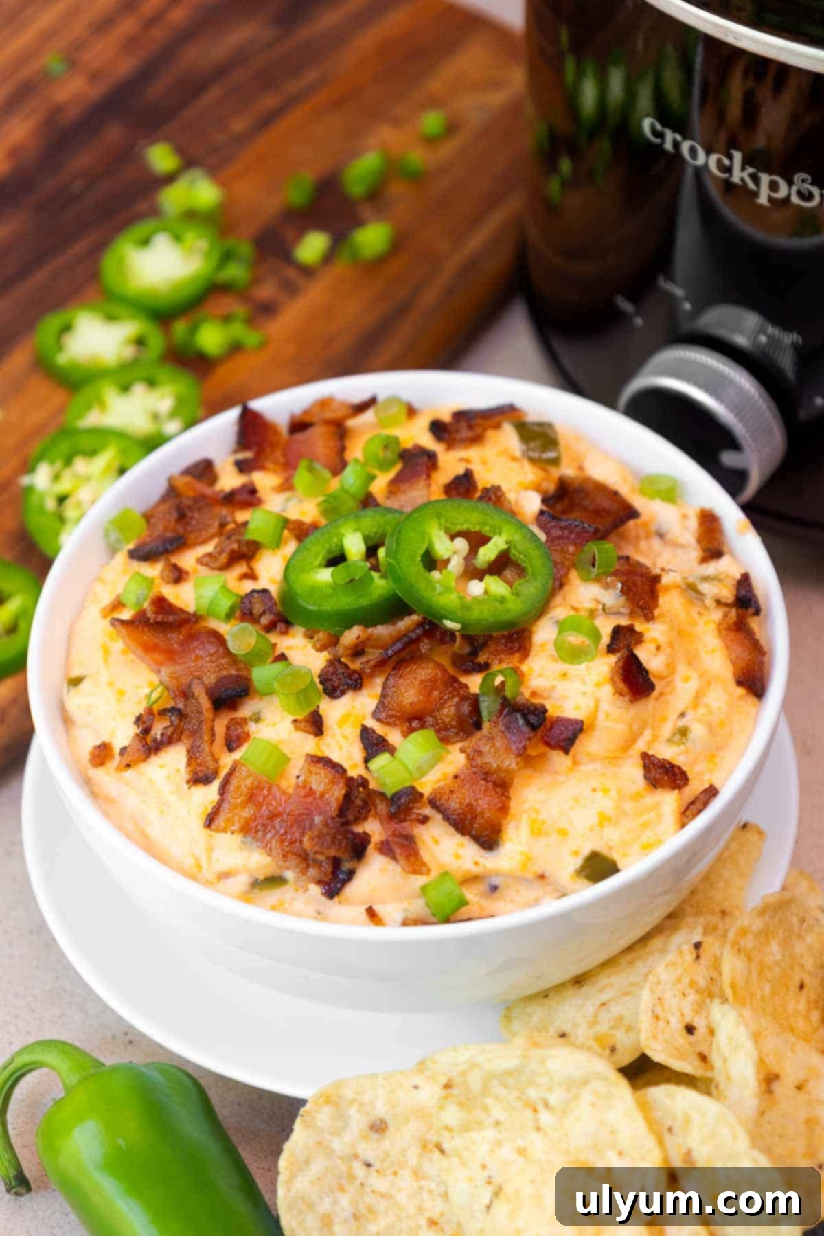 A bowl of creamy crockpot jalapeño popper dip topped with crispy bacon, sliced jalapeños, and green onions, served with tortilla chips beside a slow cooker.