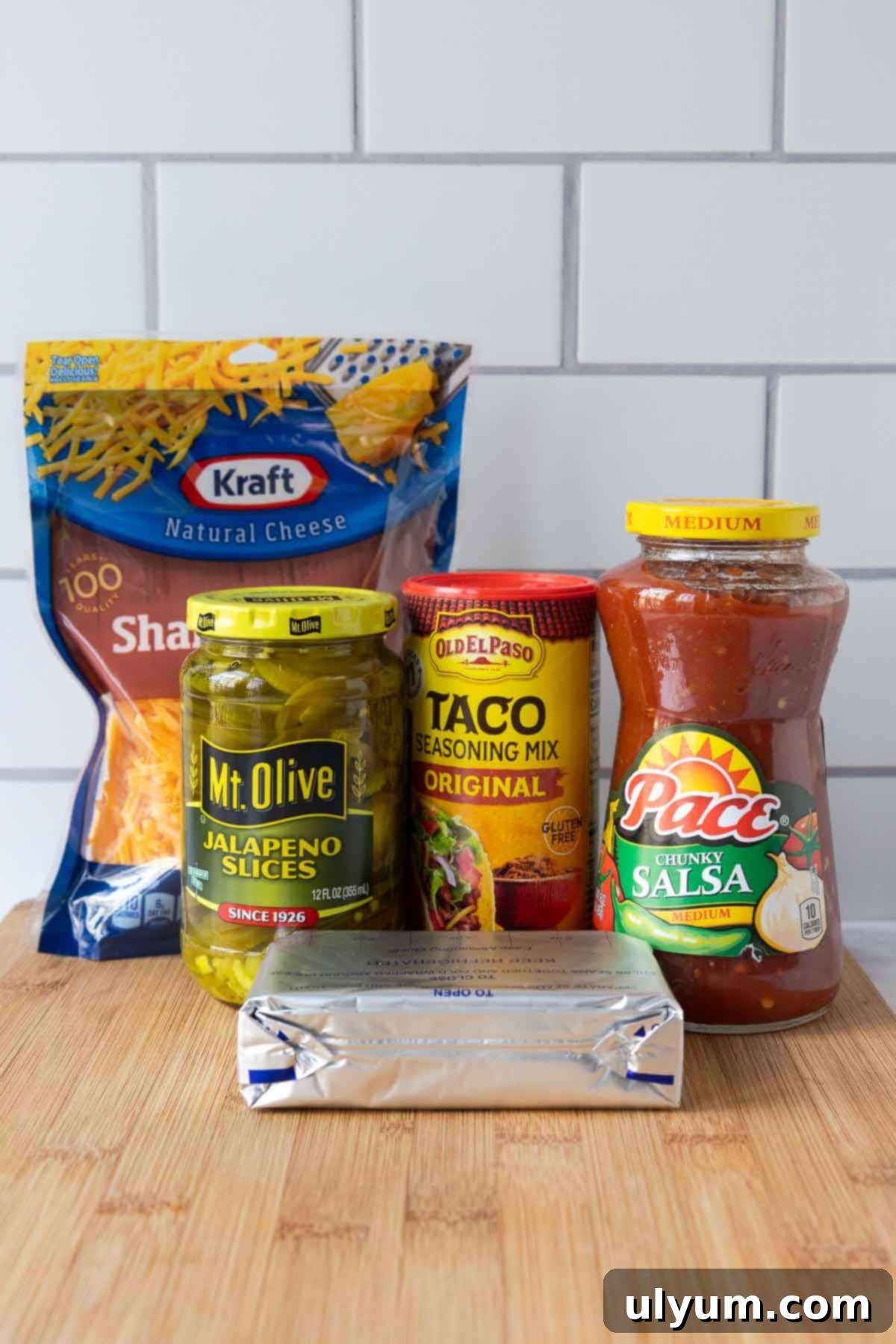 The essential ingredients for cream cheese salsa dip arranged on a wooden board: cheddar cheese, jalapeños, taco seasoning, salsa, and cream cheese.