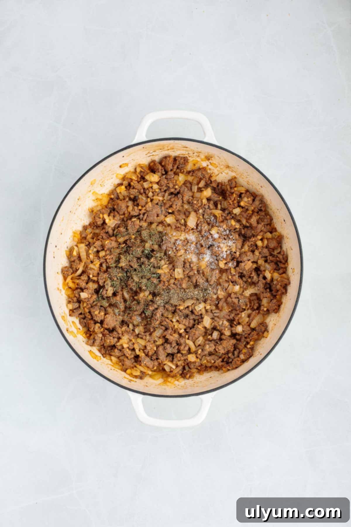 Ground beef and onions cooking in a Dutch oven with dried herbs and seasoning sprinkled over the meat.