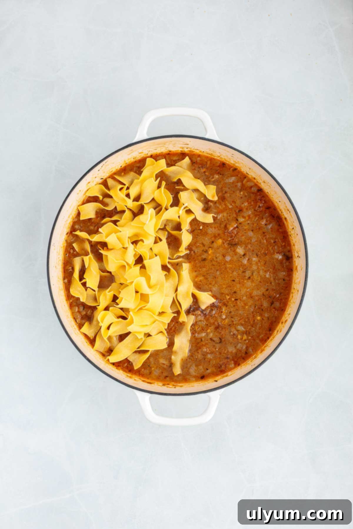 Egg noodles added to a simmering beef sauce in a Dutch oven. The uncooked noodles rest on top of the liquid before being stirred in.