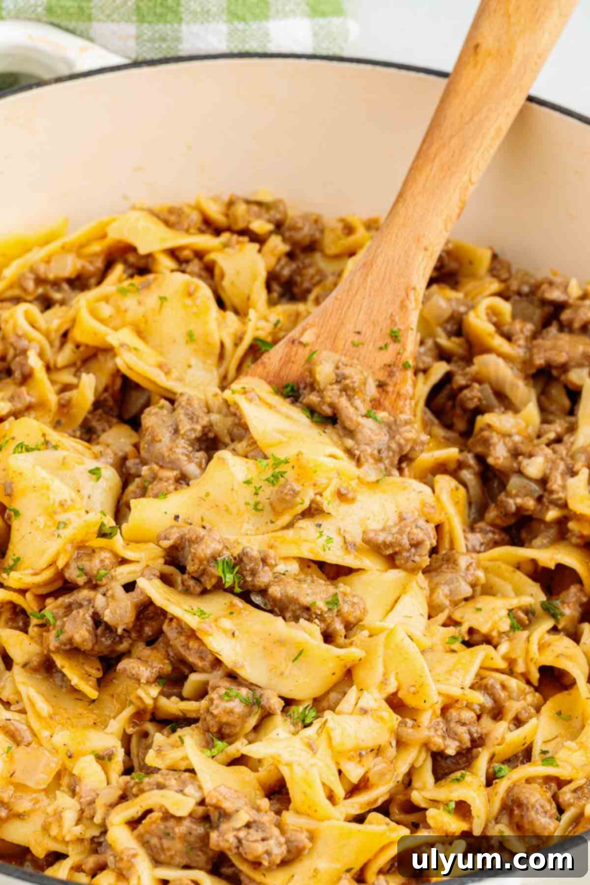 Ground beef egg noodles being stirred inside a Dutch oven with a wooden spoon. The noodles and beef are mixed together with visible parsley and softened onions.